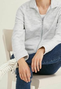 Světle šedá fleecová bunda na zip s jemnou mramorovanou texturou, nošená s modrými džínami a bílými vysokými teniskami, sedící na židli.