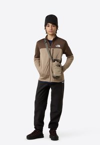 Jachetă din fleece maro și bej, cu buzunar cu fermoar pe piept, asortată cu pantaloni și cizme negre. O geantă mică bej este atașată pentru a transporta o sticlă cu apă.