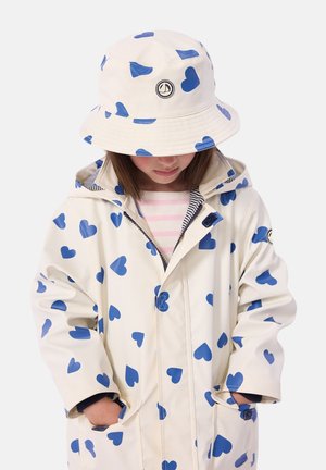 Imperméable blanc avec des motifs de cœurs bleus, doté d'une capuche, de poches frontales et d'une fermeture à boutons. Associé à un chapeau bob assorti.