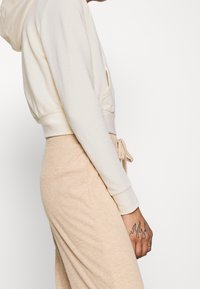 Vista laterale di una persona che indossa un hoodie crema e pantaloni a coste beige, che mostra un tatuaggio di fiamma sulla mano su sfondo bianco.
