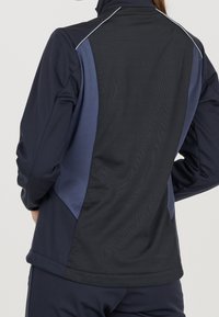 Giacca sportiva navy con un pannello contrastante di colore blu scuro, texture liscia, colletto alto e dettagli riflettenti lungo le cuciture.