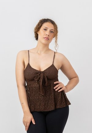 Femme aux cheveux bouclés portant un haut sans manches en dentelle marron avec un nœud à l'avant, posant avec une main sur la hanche sur un fond uni.