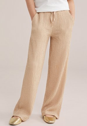 Pantaloni bej texturați cu picioare largi, talie elastică și șnur, purtați cu pantofi aurii cu talpă plată și un tricou alb, mâinile în buzunare.