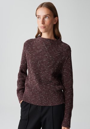 Femme avec des cheveux bruns raides portant un pull en tricot côtelé maroon à motifs et un pantalon noir, regardant légèrement vers la gauche avec les mains dans les poches.