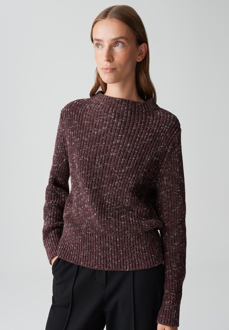 Frau mit glattem braunem Haar, die einen kastanienbraun gesprenkelten gerippten Strickpullover und schwarze Hosen trägt, leicht nach links schaut und die Hände in den Taschen hat.