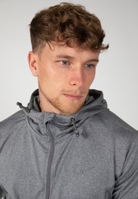 Grijze zip-up hoodie met een textuurafwerking, verstelbare trekkoorden en een subtiele logo op de borst. Heeft een hoge kraag.