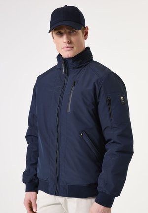 Chaqueta azul marino con cuello alto, cierre frontal con cremallera, bolsillos laterales y puños acanalados. Presenta un tejido texturizado y detalles de bolsillos laterales con cremallera.