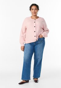 Gaiši rozā ribots kardigans ar melnām pogām, kombinēts ar plaša stila zilām džinsa biksēm un tumšām plakankāju kurpēm, stāvot pret baltu fonu.