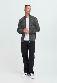 Hombre de pie que lleva un suéter gris con cremallera, camiseta blanca, pantalones negros y zapatillas blancas con rayas negras, con las manos entrelazadas al frente.