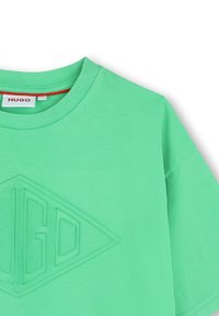 Camiseta de algodón verde menta con cuello redondeado, manga corta y un logo triangular elevado con el texto "HUGO" en el centro.
