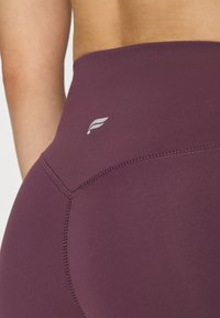 Bordo tajice visokog struka izrađene od rastezljivog, glatkog materijala s malim bijelim logotipom i ravnim šavovima za dodatni detalj.