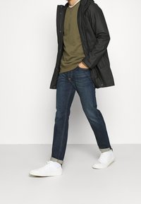 Uomo che cammina, indossando jeans blu scuri risvoltati alle caviglie, sneakers bianche, un maglione verde oliva e un impermeabile nero con una mano in tasca.