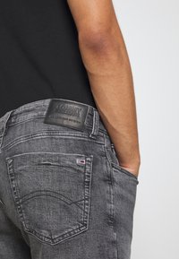 Jeans in denim grigio scuro con una toppa in pelle etichettata "TOMMY JEANS." Caratteristiche includono tasche posteriori curve e un piccolo logo in rosso, bianco e blu.