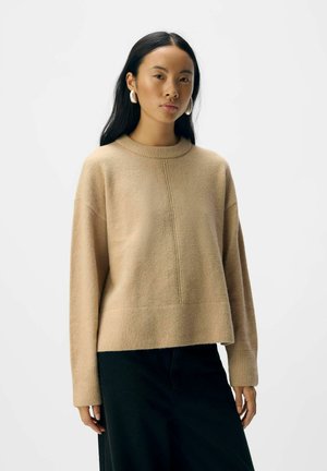 Beige knit sweater with a crew neck, dropped shoulders, and ribbed cuffs. Features a central seam and a relaxed fit. Paired with black pants.