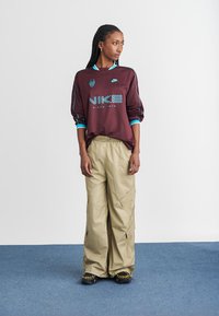 Burgundijas garā piedurkne Nike džersija ar tirkīza akcentiem; brīvās beigu krāsas cargo bikses; melnas un dzeltenas sporta apavi; minimālistisks fons.