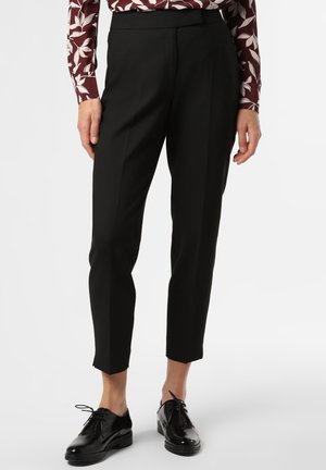 Vrouw draagt zwarte op maat gemaakte enkelhoge broek, bloemenblouse met lange mouwen en zwarte veterschoenen in Oxford-stijl, staand tegen een witte achtergrond.