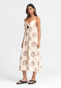 Maksi haljina od krem tkanine s smeškim cvjetnim uzorkom. Ima čvor na prednjem dijelu i tanke naramenice. U kombinaciji s crnim japankama.