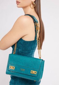 Teal läderhandväska med en texturerad finish, guldfärgad hårdvara och en kedjestropp. Har en logotyp från varumärket och en strukturerad form.