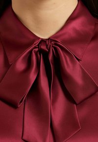 Blusa di raso bordeaux con un ampio collarino a fiocco, caratterizzata da una texture liscia e un design strutturato, che enfatizza un'elegante caduta e lucentezza.