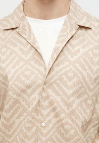 Μπεζ υφασμάτινο πουκάμισο με κουμπιά, με αφηρημένο γεωμετρικό σχέδιο, φορεμένο πάνω από λευκή ριμπ πλεκτή μπλούζα με στρογγυλή λαιμόκοψη.