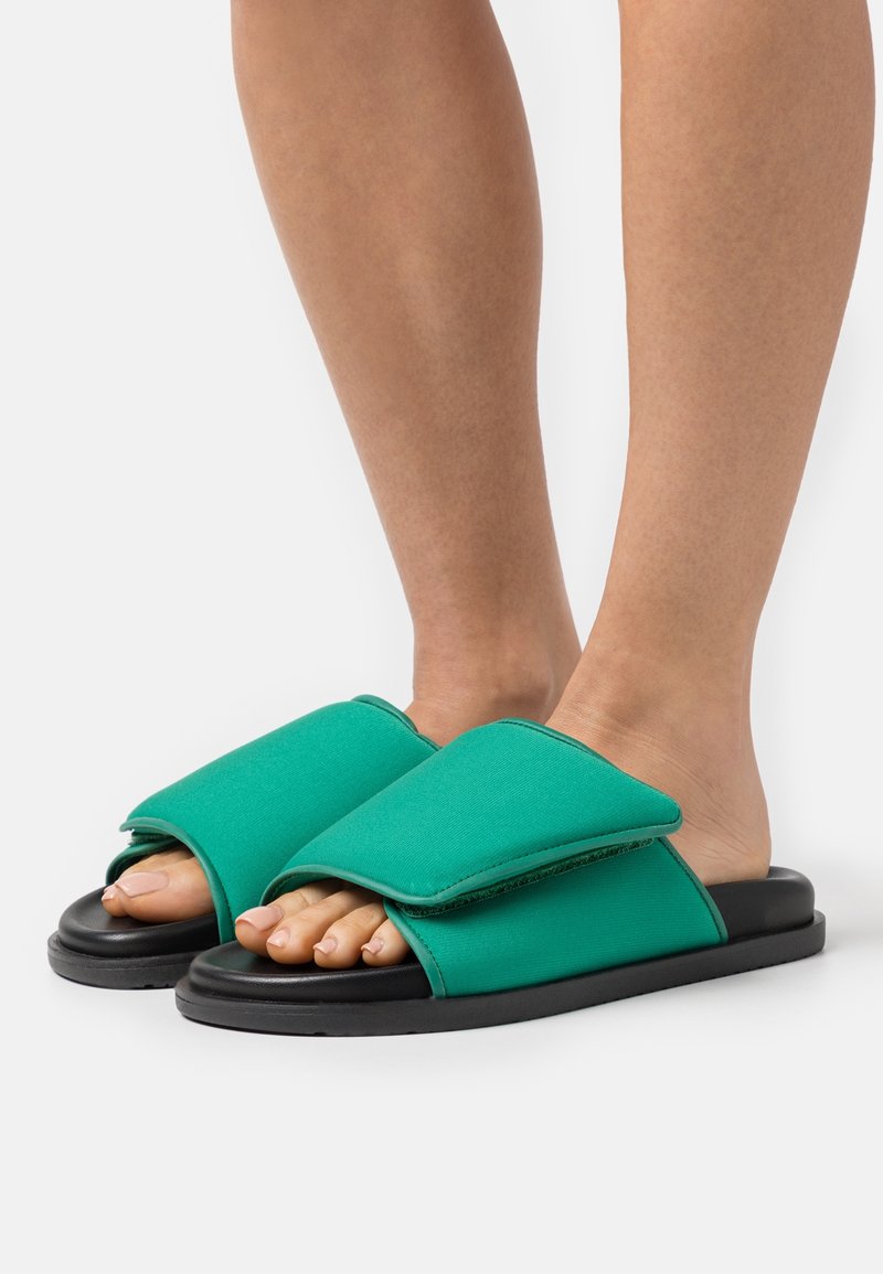 Pieds portant des sandales slide en tissu vert avec de larges brides réglables et des semelles noires sur fond blanc.