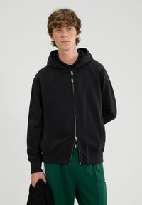 Felpa nera con zip realizzata in morbido tessuto, dotata di cappuccio, con un logo discreto, polsini a coste e una vestibilità comoda, abbinata a pantaloni verdi.