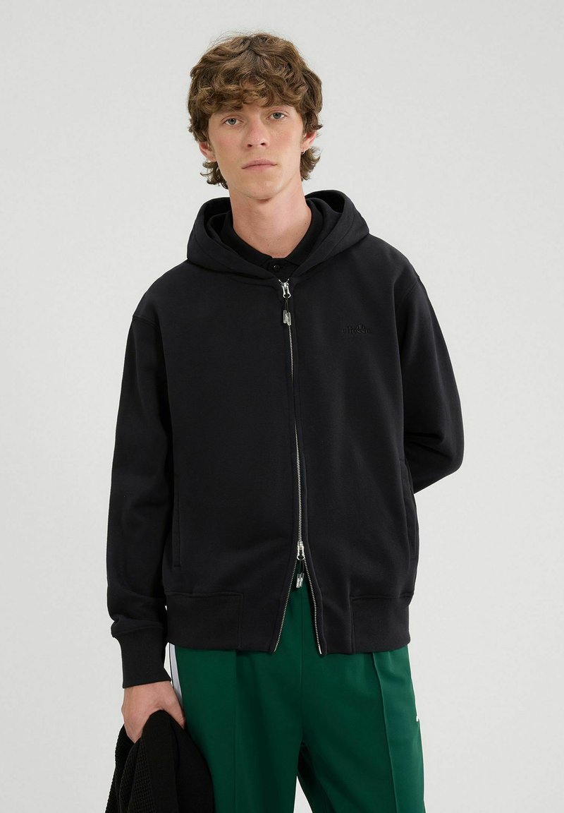 Felpa nera con zip realizzata in morbido tessuto, dotata di cappuccio, con un logo discreto, polsini a coste e una vestibilità comoda, abbinata a pantaloni verdi.