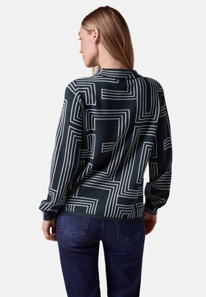 Svart tröja med ett geometriskt vitt linjemönster; har rund hals och långa ärmar, matchat med blå denimjeans.