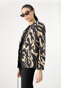 Blazeren har et abstrakt mønster i sort og beige, enkelt knaplukning og en afslappet pasform. Båret med en sort top og mørke jeans.