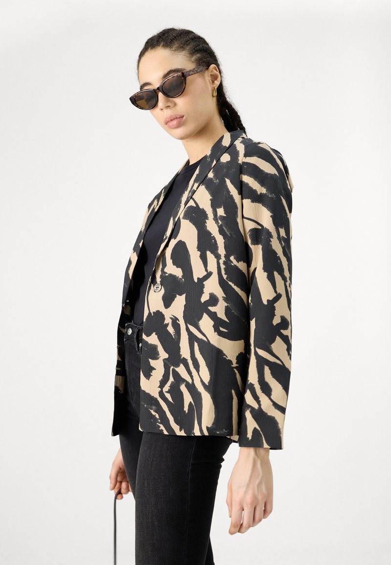 Le blazer présente un motif abstrait noir et beige, une fermeture à un bouton et une coupe décontractée. Porté avec un haut noir et un jean foncé.