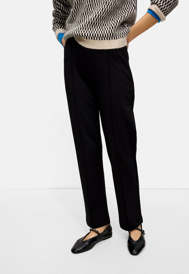 Pantalons noirs ajustés avec une coupe droite, présentant une texture lisse et des détails de couture visibles. Associés à des chaussures plates noires.