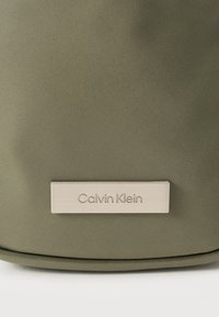 Πράσινη τσάντα από νάιλον με ασημένια ορθογώνια πινακίδα λογότυπου με χαραγμένο το όνομα «Calvin Klein», με υφή στην επιφάνεια και στρογγυλεμένες γραμμές.