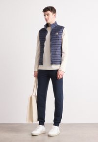 Vesta puffer de culoare bleumarin peste un pulover bej, asortată cu pantaloni închiși și adidași albi. Modelul ține o geantă tote bej. Textură netedă, design minimalist.