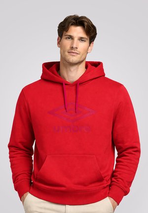 Mężczyzna o brązowych włosach, ubrany w czerwoną bluzę Umbro z kapturem i przednią kieszenią, stoi z rękami w kieszeniach na tle jednolitego, jasno szarego tła.