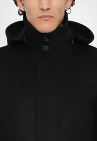 Czarny wełniany płaszcz z wysokim kołnierzem, z dwoma guzikami i ukrytym zamkiem błyskawicznym. Tkanina ma gładką teksturę i wyrazisty design.