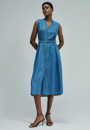 Mujer con vestido midi de denim azul sin mangas, con botones delanteros y cinturón, combinado con sandalias marrones de tacón alto, de pie sobre un fondo liso.