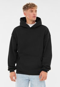 Sudadera negra de gran tamaño, hecha de algodón, con un bolsillo frontal, puños acanalados y capucha con cordón, presentada con jeans azul claro.