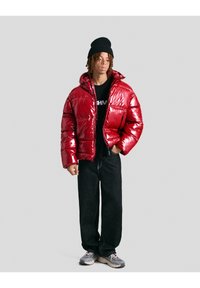 Giacca a vento rossa con finitura lucida, chiusura con zip sul davanti e cappuccio, abbinata a pantaloni neri e sneakers grigie. Berretto a maglia nero sulla testa.