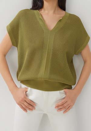 Frau trägt olivgrünes, kurzärmeliges Stricktop mit V-Ausschnitt und strukturiertem Muster, kombiniert mit weißer Hose, Hände auf den Hüften ruhend.