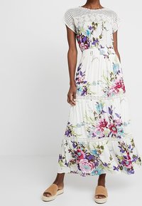 Maxi kjole i let hvidt stof med blomstermønstre i pink, lilla og blå, med spidse detaljer ved halslinjen og lagdelte flæser.