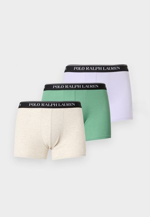 Muskularny mężczyzna stoi boso, nosząc białe bokserki z czarnym pasem z logo Polo Ralph Lauren na jednolitym jasnym tle.