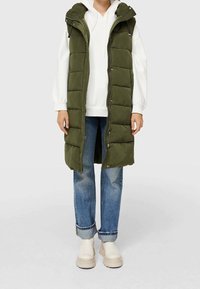 Olivgrüne gesteppte lange Weste mit hohem Kragen und Reißverschluss, über einem weißen Hoodie getragen, kombiniert mit hochgekrempeltem Denim und beigen Stiefeln.