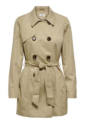 Kvinde iført en beige trenchcoat med bælte over en blå sweater, kombineret med blå jeans og sorte sko, står med den ene hånd i lommen.