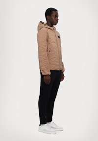 Beige quiltet jakke med hætte, med et sort mærke på brystet, båret med sorte bukser og hvide sneakers.