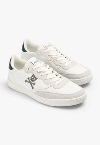 Adidași din piele albă cu accente din suede gri, având un logo cu craniu și oase încrucișate pe lateral. Vârf rotunjit și talpă din cauciuc texturată.