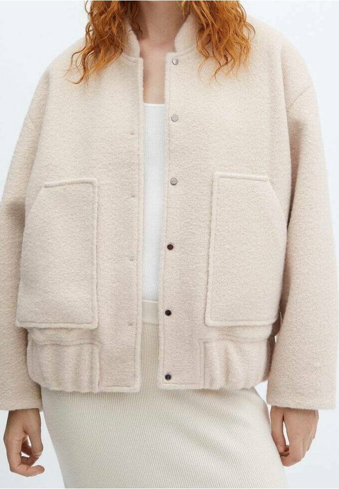 Femme portant une veste en laine beige avec de grandes poches avant et des boutons-pression sur un top côtelé blanc et une jupe.