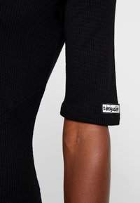 Gros plan sur le bras d'une personne portant une chemise noire texturée à manches courtes avec une étiquette tissée portant l'inscription "#desigual.com" près du poignet.