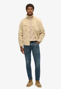 Chaqueta de algodón beige sobre un suéter claro, combinada con unos vaqueros azul oscuro y botas beige. Cuenta con dos bolsillos en el pecho y botones.