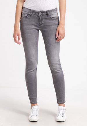Frau trägt slim-fit ausgewaschene graue Jeans, ein weißes Oberteil und weiße Schnürsneaker, steht vor schlichthintergrund.