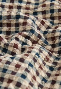 Tessuto con motivo a scacchi in crema, blu navy e bordeaux. Il materiale presenta una texture con dettagli intrecciati e un aspetto morbido, leggermente accartocciato.